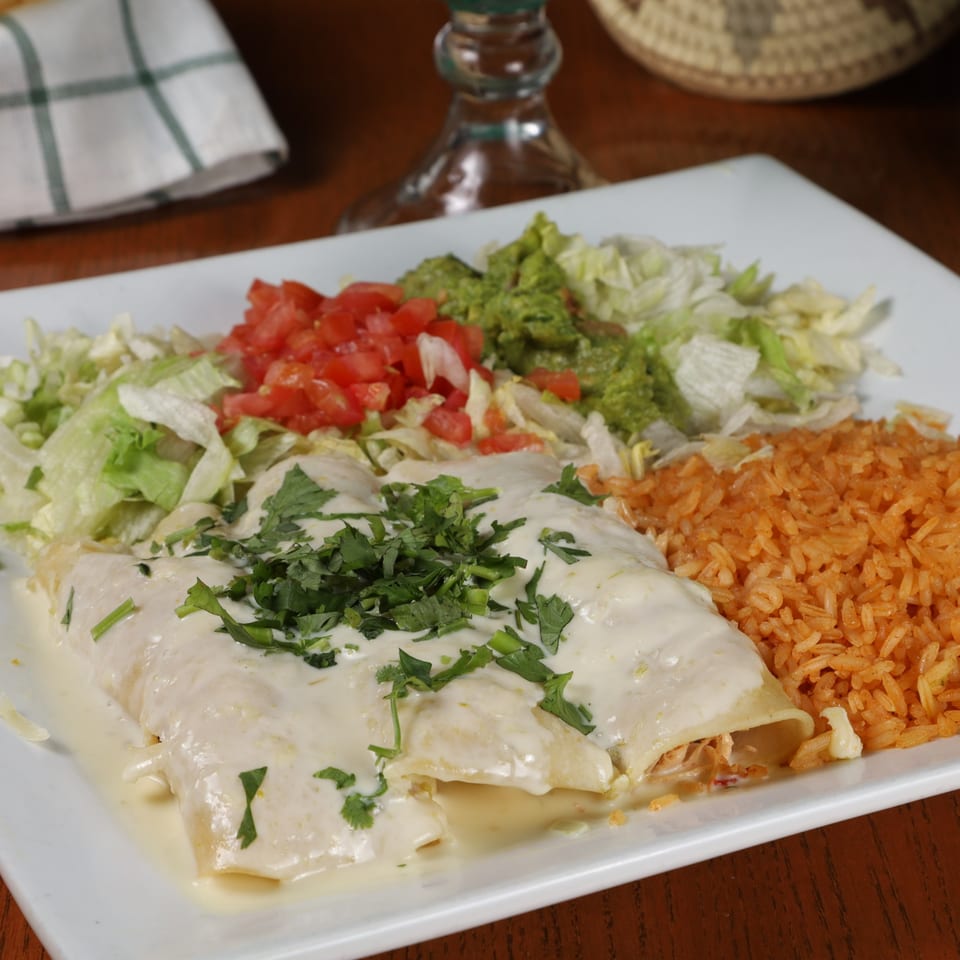 Creamy enchiladas with rice, lettuce, tomatoes, and guacamole on a white plate.