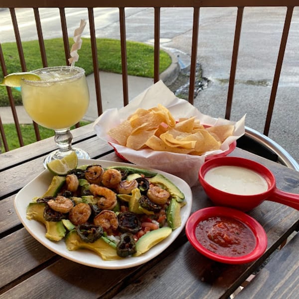 Shrimp and avocado dish with chips, salsa, queso, and a margarita on a wooden table.