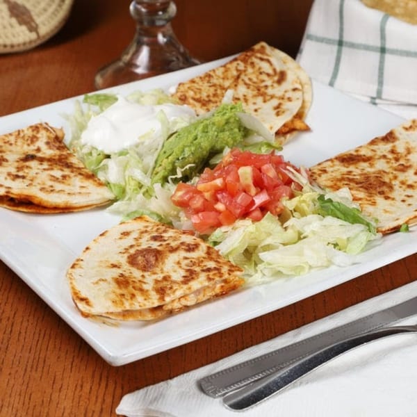 Four quesadilla wedges with shredded lettuce, diced tomatoes, guacamole, and sour cream on a white plate.