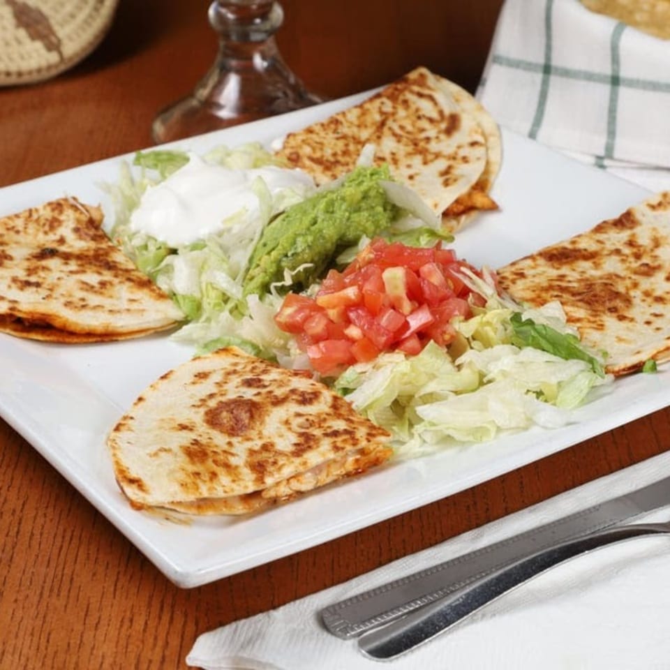 Four quesadilla wedges with shredded lettuce, diced tomatoes, guacamole, and sour cream on a white plate.