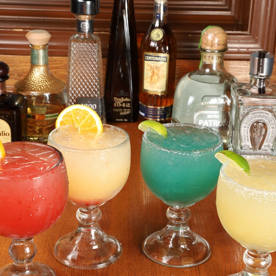 Four colorful margaritas in goblet glasses with tequila bottles in the background at La Fuente Mexican Bar & Grill.