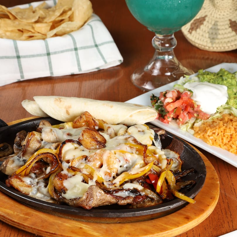 Sizzling fajitas with melted cheese, bell peppers, and onions, served with tortillas, rice, and guacamole.